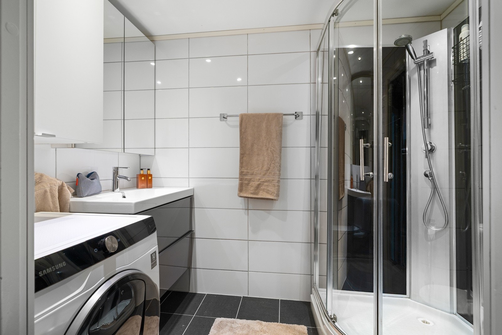 Flislagt baderom med varmekabler i gulv og dusjkabinett. Badet har en nyere servant, med en mørk innredning med speilskap, belysning samt opplegg for vaskemaskin Galleribilde