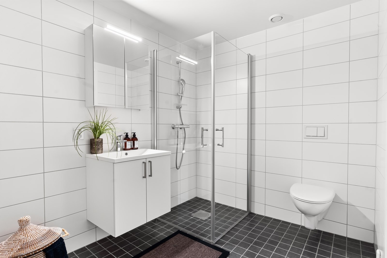 På badet er det baderomsinnredning med nedfelt servant m/ettgreps servantbatteri, speil med integrert belysning. Videre er det ettgreps dusjbatteri med dusjgarnityr (fleksibel slange og hånddusj) og dusjhjørne. Vegghengt klosett i hvitt porselen. Galleribilde