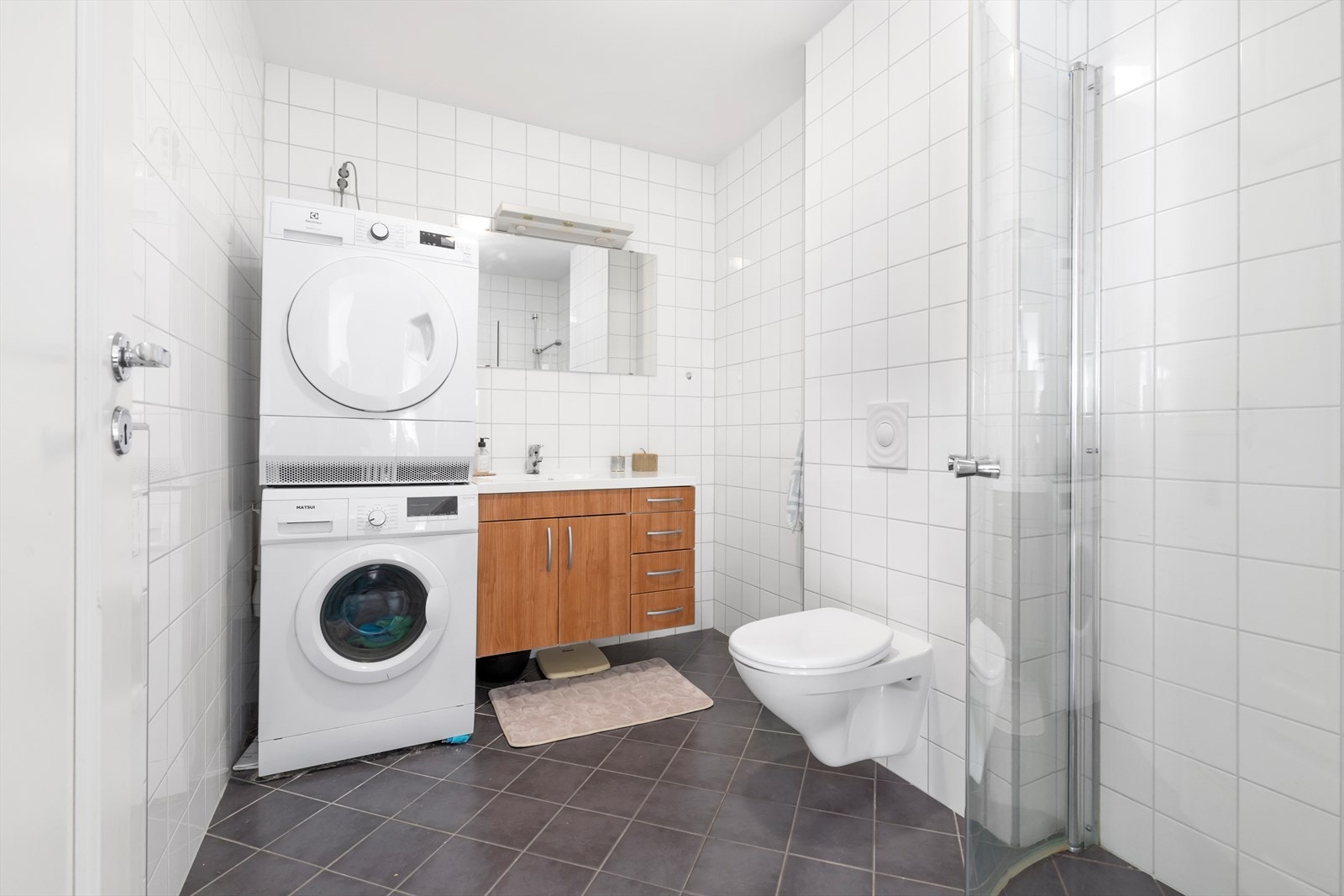 Bad/vaskerom har flislagte vegger og flislagte gulv med vannbåren varme | rommet er utstyrt med dusjhjørne med buede glassdører, vegghengt wc, baderomsinnredning og opplegg for vaskemaskin og tørketrommel Galleribilde