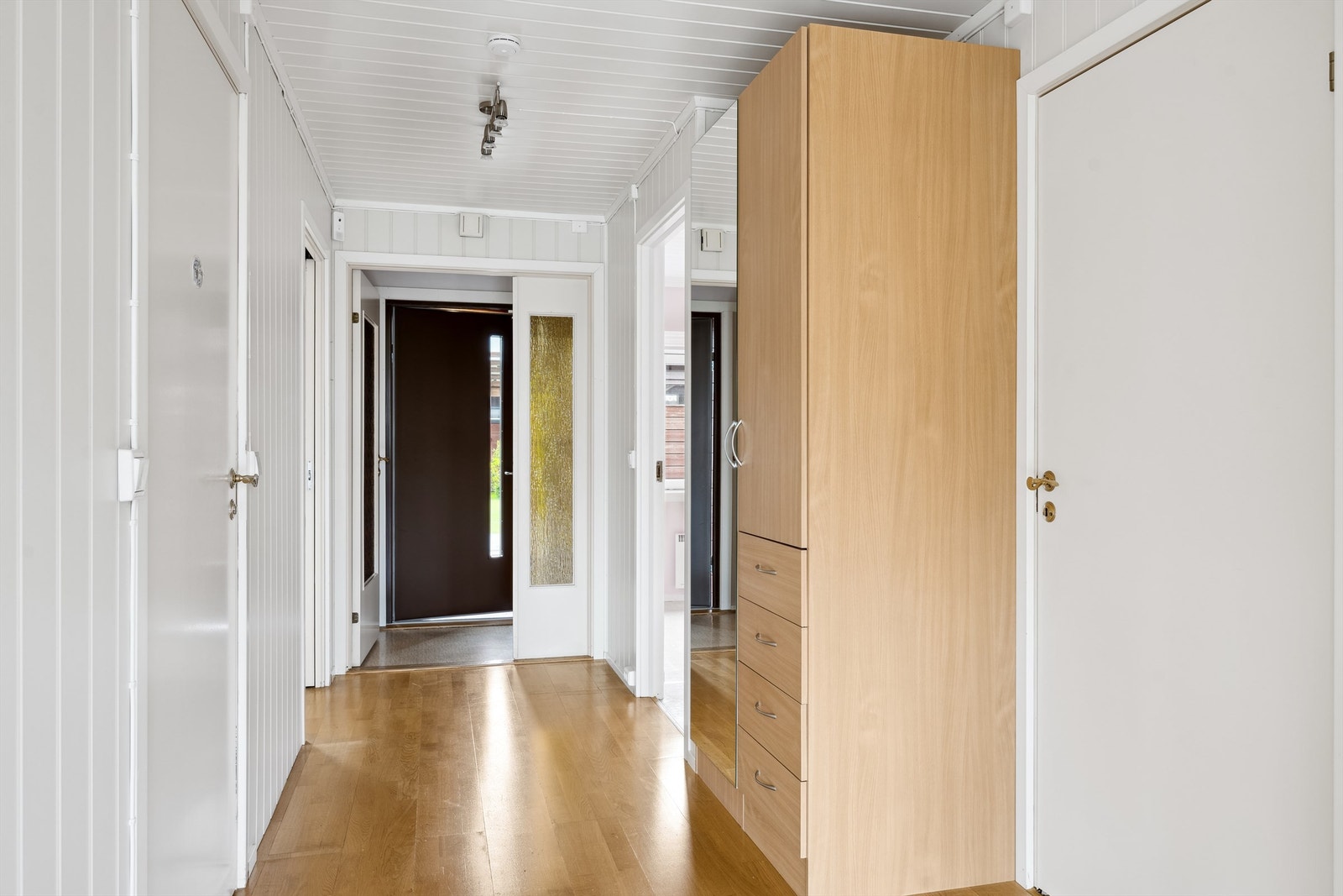 Her kommer du inn i et lite vindfang med belegg på gulvet og en dør med glassfelt som fører deg videre inn i gangen.
Gangen er lysmalt og smal med adkomst til alle rommene i leiligheten. Galleribilde
