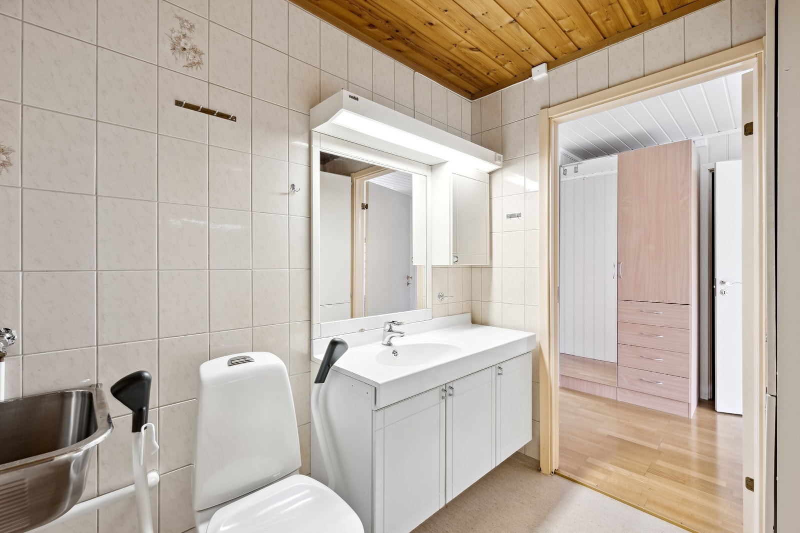 Badet er innredet med servant på hvit skapinnredning, vegghengte skap, speil, gulvmontert toalett, dusjhjørne med dusjvegger og armatur montert på vegg, utslagsvask montert på veggen og opplegg for vaskemaskin. Galleribilde
