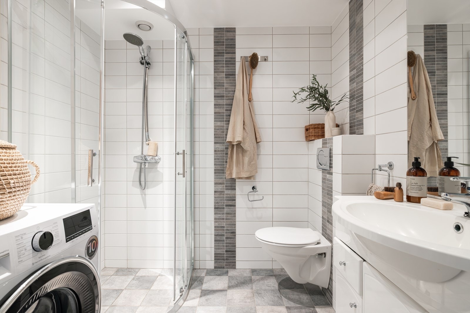 Lyst og delikat bad innredet med fliser på gulv og vegger i tidsriktige farger. Rommet er utstyrt med vegghengt toalett, dusjhjørne med glassdører, servant med underskap og speil med integrert belysning. Galleribilde