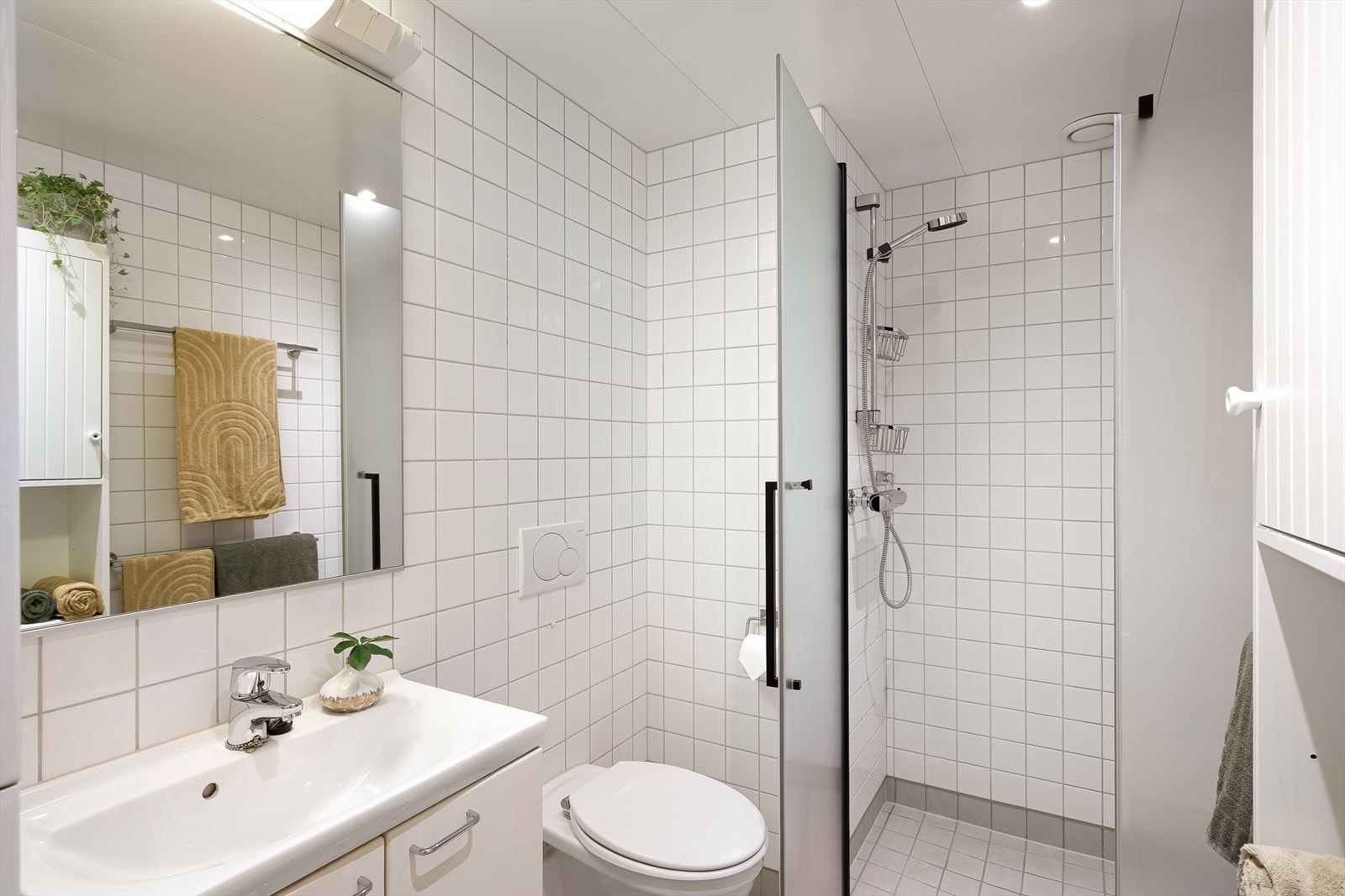 Pent helfliset baderom utstyrt med hvit servant, veggmontert toalett, og dusjhjørne med glassdør. Gulvvarme Galleribilde