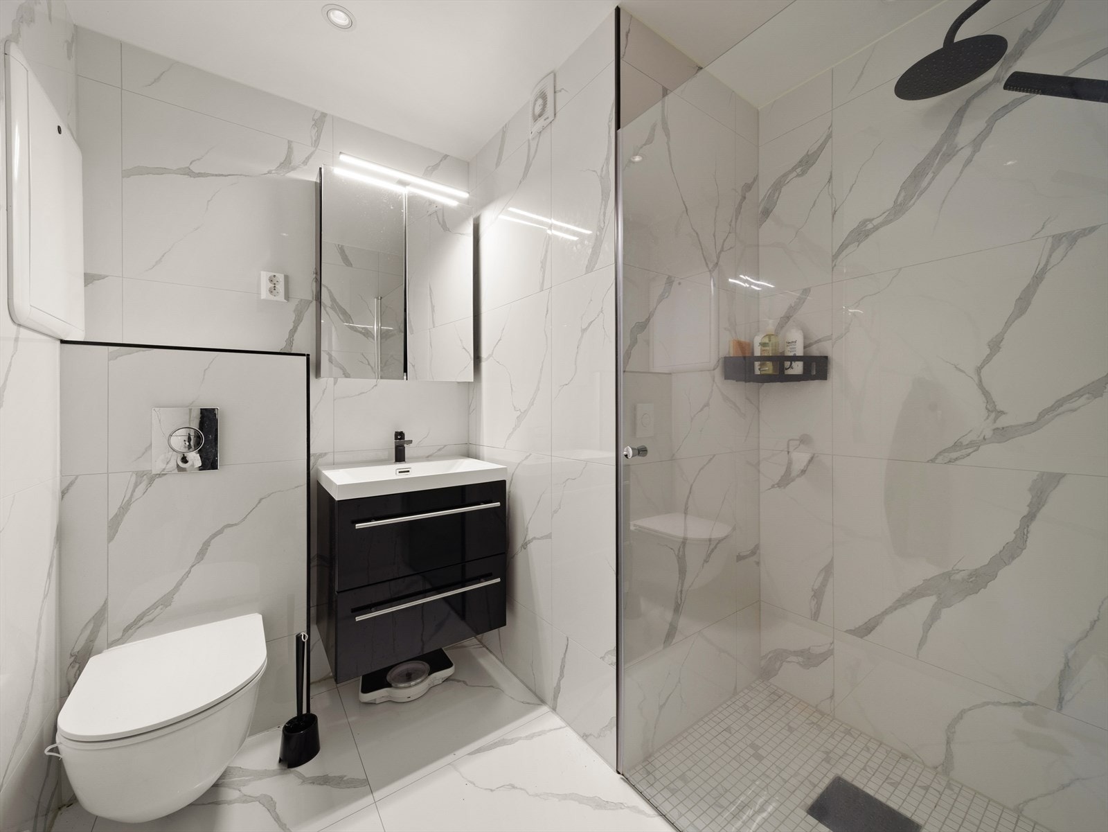 Badet er pent innredet med nedsenket dusjsone med glassvegg og glassdør, vegghengt klosett med innebygd sisterne og møblement med servant Galleribilde