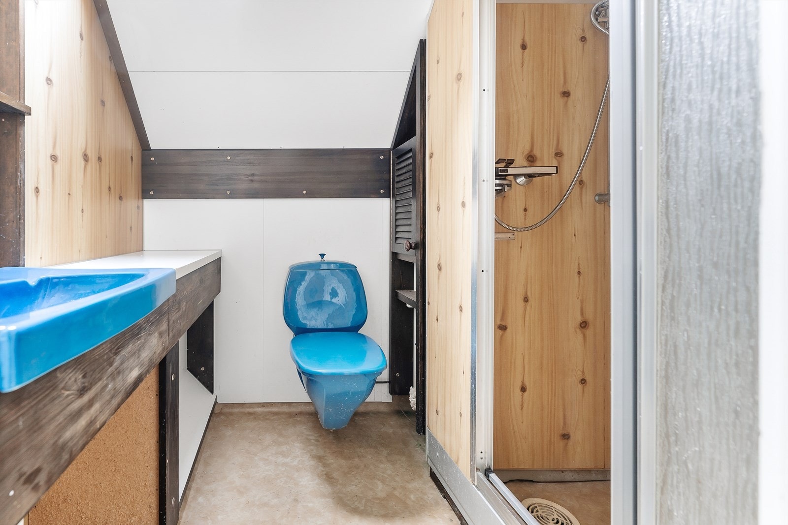 Bad på loft fra ca. 1980 med våtromsbelegg på gulv og våtromsplater på vegger, malt himling, dusjnisje med dusjing på vegg/våtromsplater, et istykkerfrosset gulvtoalett og varmekabel i gulvet. Galleribilde