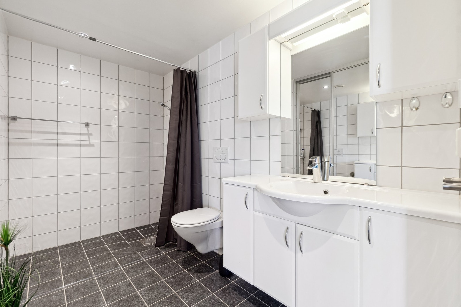 Badet fremstår tidløst takket være en lys og mørk utførelse på overflater og innredning. Her er det vegghengt toalett, dusjsone med garnityr på vegg, god oppbevaringsplass i baderomsinnredningen, og opplegg for vaskemaskin. Galleribilde