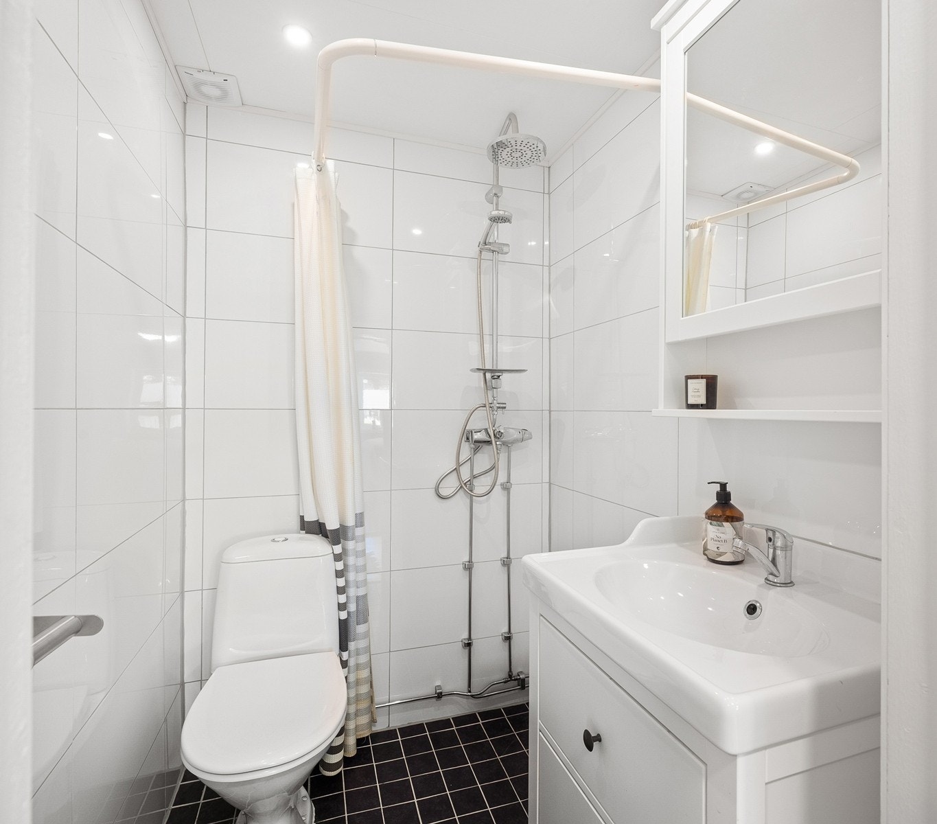 Badet har et moderne uttrykk med hvite veggfliser og kontrastfulle sorte gulvfliser med gulvvarme. Innredningen består av et underskap/overskap, servant, speil og dusjsonen har regnfallsdusj. Galleribilde