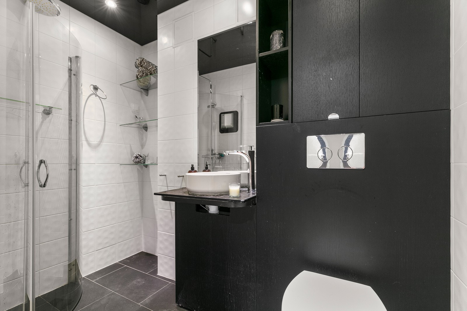 Boligen inneholder et stilfullt bad og et separat vaskerom. Badet har mørke fliser på gulv, hvite fliser på veggene, samt sort stretch ceiling med glass- og speileffekt og downlights, som gir rommet et romslig og moderne preg. Galleribilde