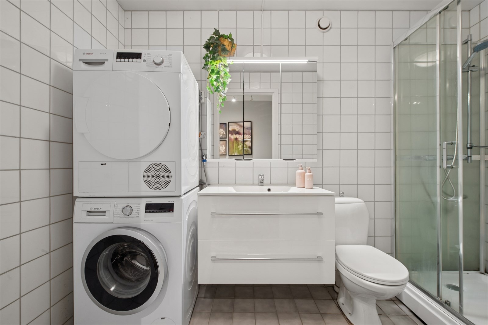 Tidløst flislagt baderom med varmekabler på gulv. Rommet har innredning med nedfelt servant, toalett, dusjkabinett, opplegg for vaskemaskin og tørketrommel. Galleribilde