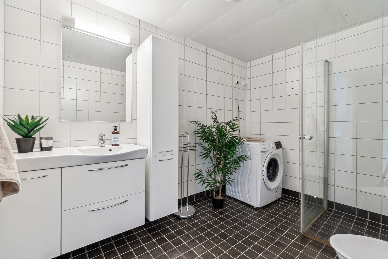 Ved gangen finner du boligens bad. Badet er av god størrelse og fremstår tidløst med mørke fliser på gulv og lyse veggfliser. Det er utstyrt med bl.a. toalett, speil, dusjsone, servant med underskap, baderomsinnredning og opplegg for vaskemaskin. Galleribilde