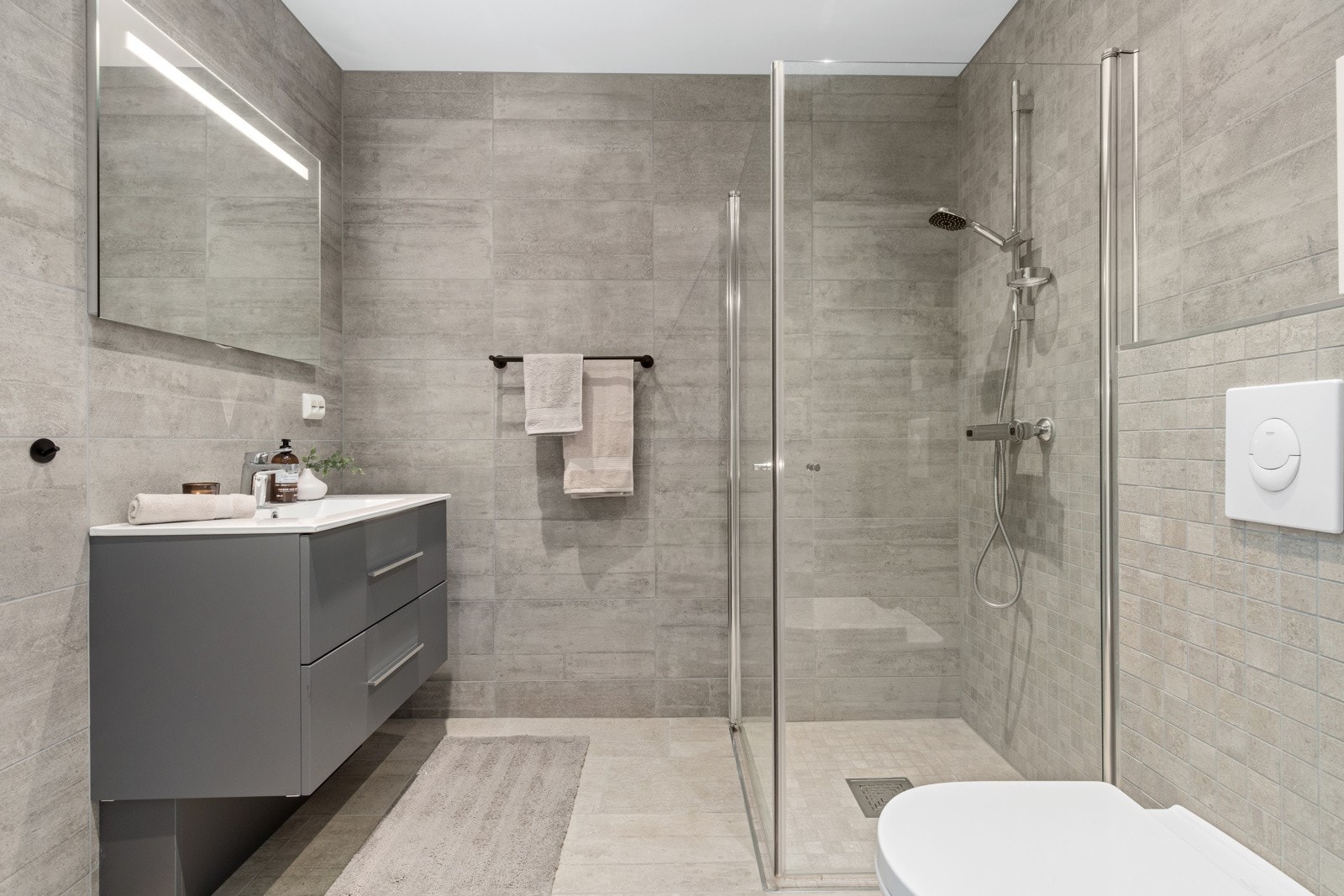 Badet er flislagt ig innbydende. Her er det montert skuffeinnredning, speil, veggmontert wc og dusjhjørne. Galleribilde