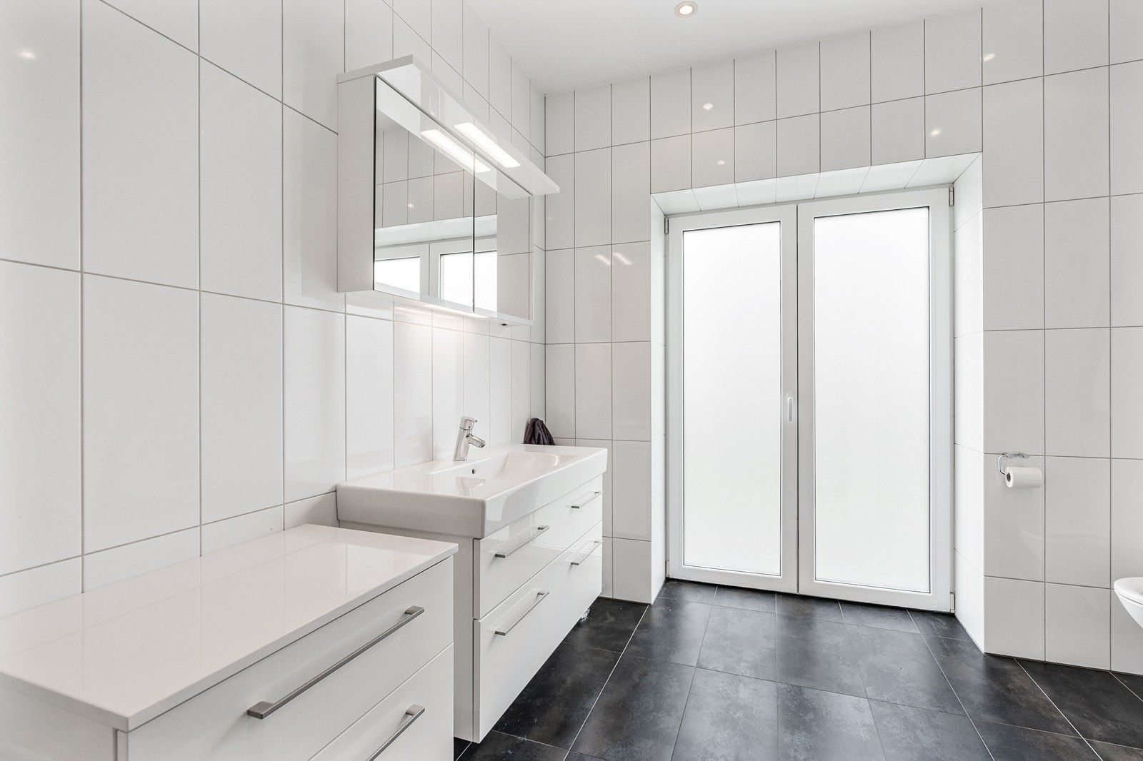 Etasjens baderom er helfliset i lekre og tidløse farger. På gulvet er det valgt en dus jordtone, mens det er store, lyse fliser på veggene. Galleribilde