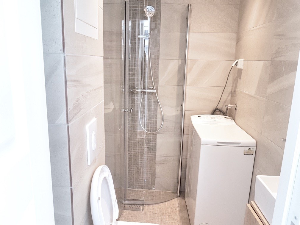 Flislagt bad med servant på underskap og speil over, gulvstående wc, dusjhjørne med innfellbare glassdører og opplegg for vaskemaskin. Galleribilde