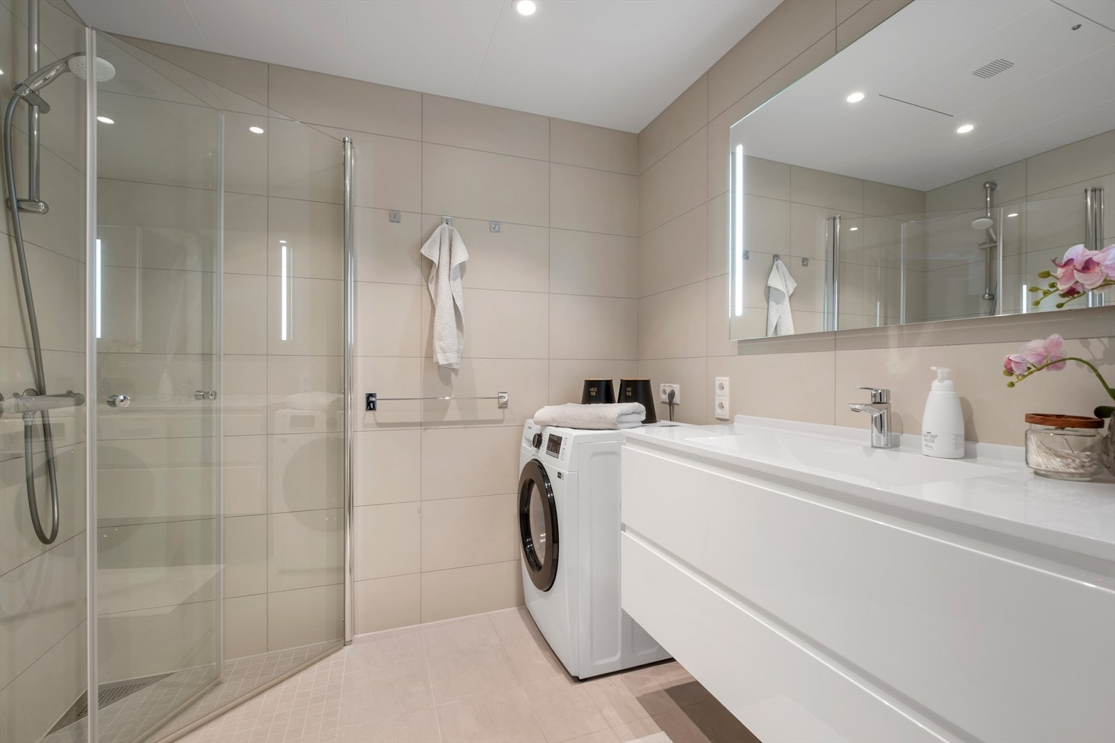 Moderne og lekkert flislagt bad i beige med behagelig gulvvarme. Badet er innredet med praktiske innfellbare dusjdører av glass og dusjgarnityr på vegg, veggmontert toalett, innredning med servant og opplegg for vaskemaskin. Galleribilde