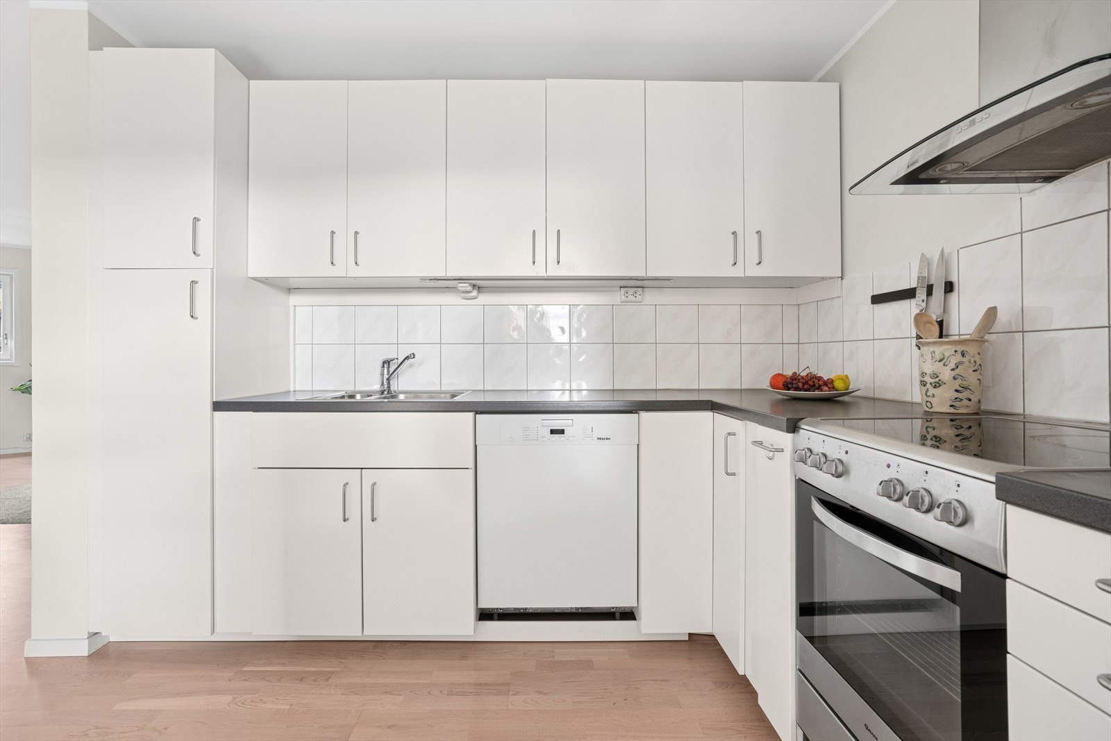Tidløs kjøkkeninnredning med hvite glatte fronter. Benkeplate i laminat med nedfelt oppvaskkum. Det er fliser mellom benk og overskap. Komfyr og oppvaskmaskin medfølger. Galleribilde