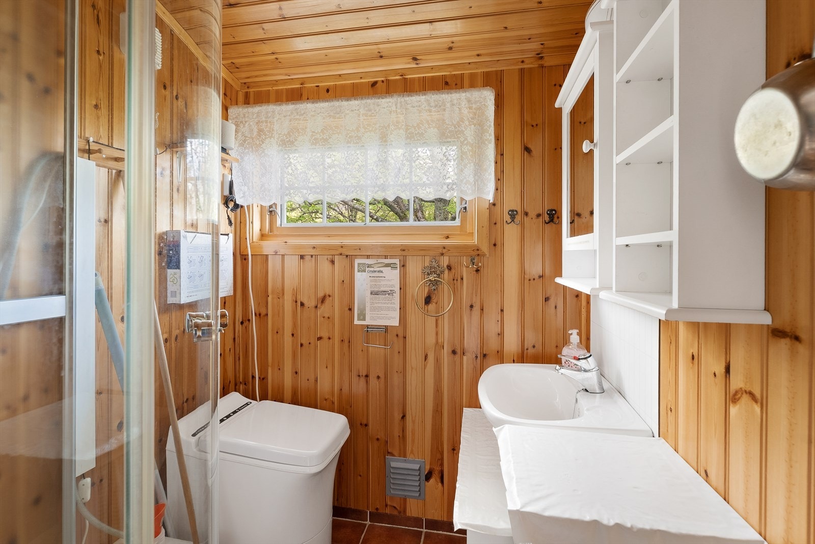 Enkelt innredet baderom med forbrenningstoalett, dusjkabinett, vask og oppbevaringsmøblement. Galleribilde