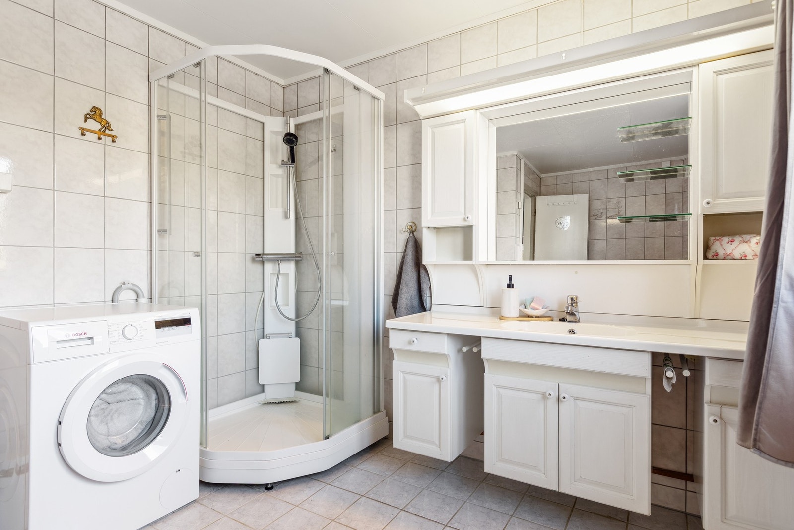 Badet er flislagt med gulvmontert toalett, dusjkabinett, opplegg for vaskemaskin og servant i innredning. Galleribilde