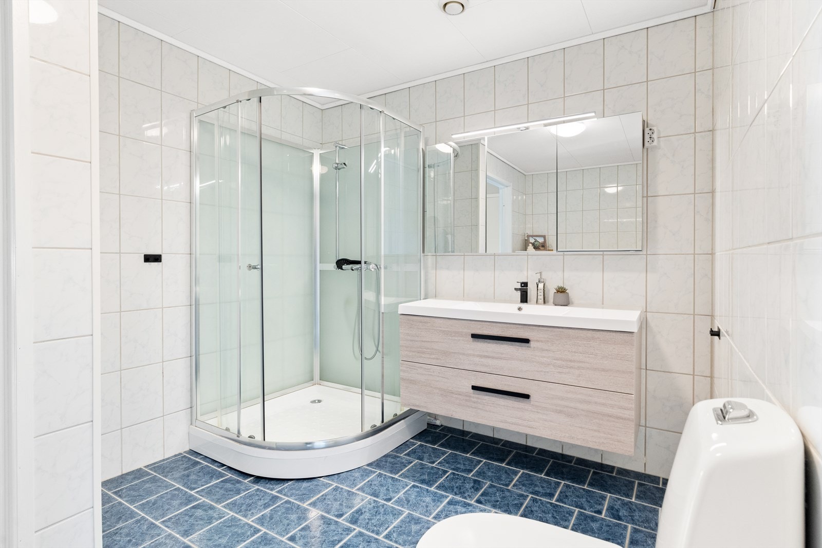 Badet er praktisk innredet med slette fronter og heldekkende servant med 1-greps blandebatteri. Speil med belysning over servant, dusjkabinett og gulvmontert toalett gjør dette til et funksjonelt og lettstelt baderom. Galleribilde