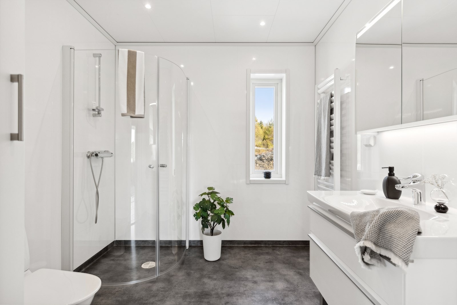 Badet er lyst og lekkert innredet med toalett, servant, dusjhjørne, håndkletørker og praktisk skap for oppbevaring. Galleribilde