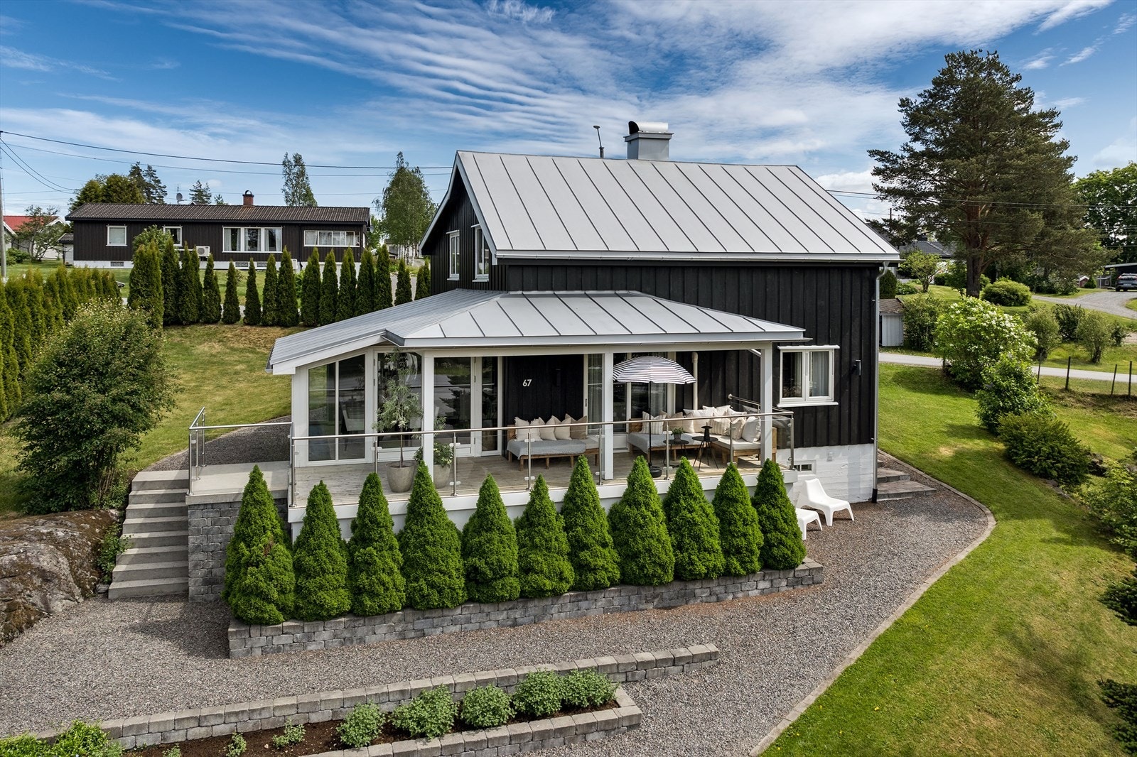 Pen fasade med tømmermannspanel, store vindusflater i påbygget deler av stuen og en flott, delvis overbygget terrasse. Galleribilde