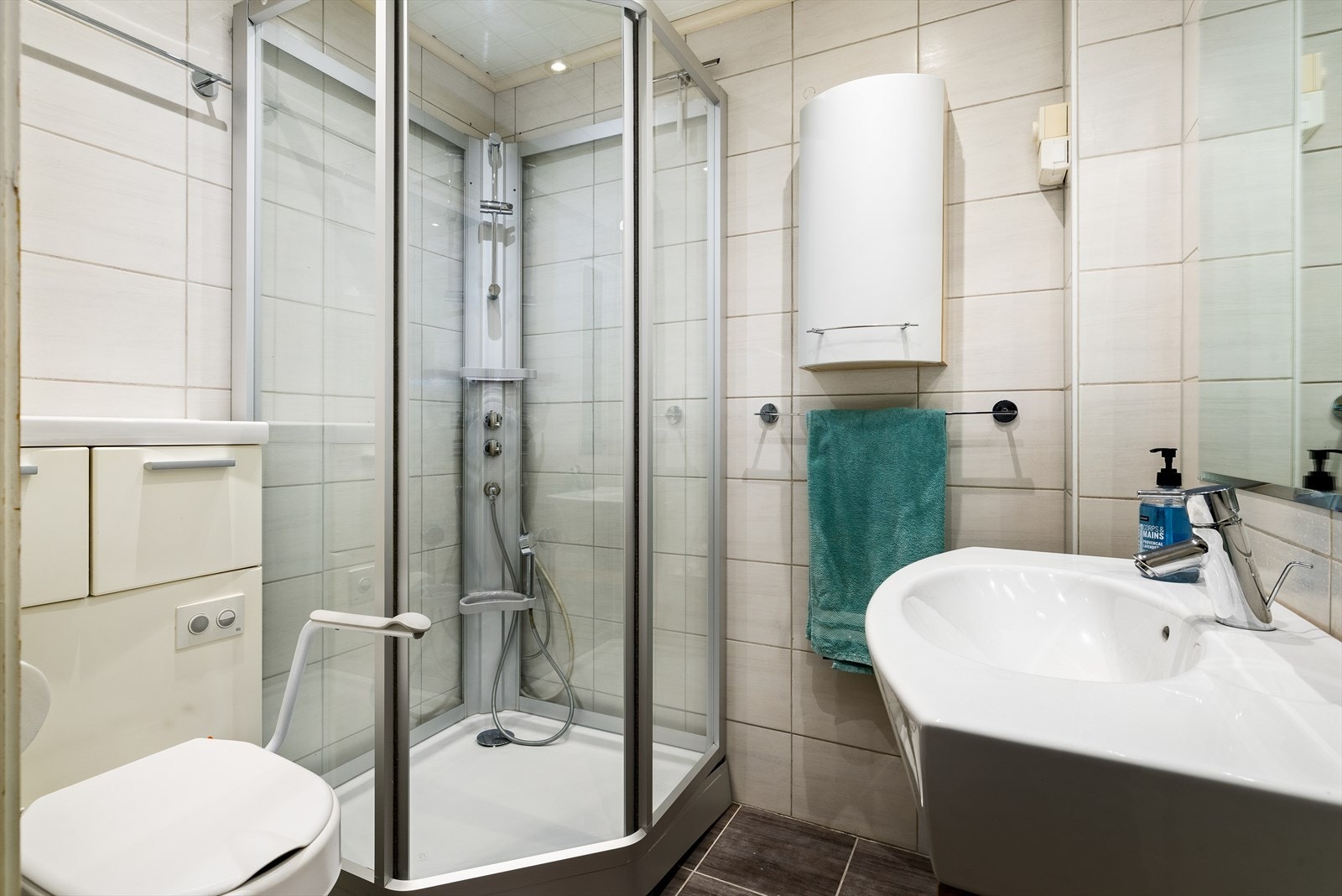 Baderommet har et stilrent uttrykk med flislagte vegger og gulv, samt elektriske varmekabler. Taket er kledd med MDF-panelbord og har innfelte downlights.
Innredet med servantskap med nedfelt servant, speil, dusjkabinett, vegghengt skap og toalett. Galleribilde