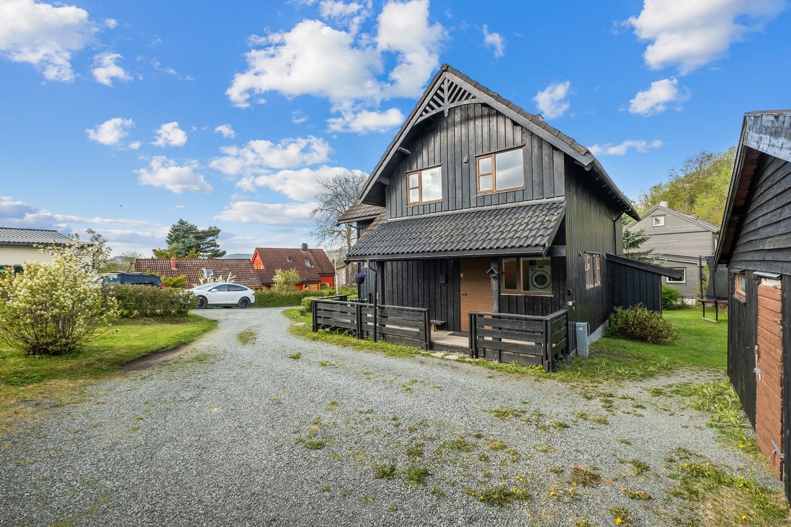 Flere oppstillingsplasser på tomten gir god plass til flere biler og el-billader er montert på husveggen. Nedgang til kjellerdel vises til høyre i bildet. Galleribilde