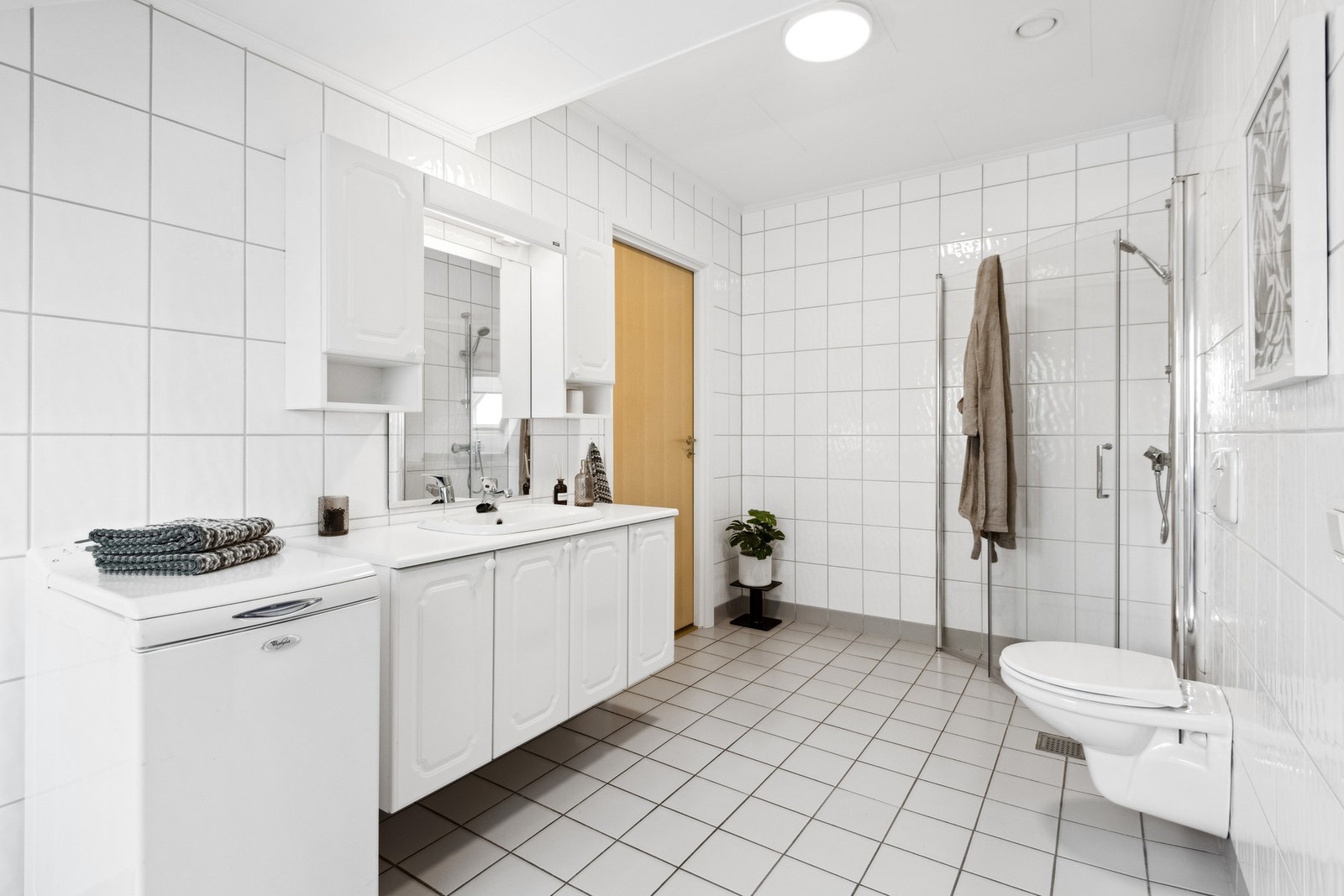 Badet har fliser på gulv og vegger. Rommet er utstyrt med åpen dusjløsning med glassdører, vegghengt toalett og servant i innredning, samt opplegg for vaskemaskin. Galleribilde