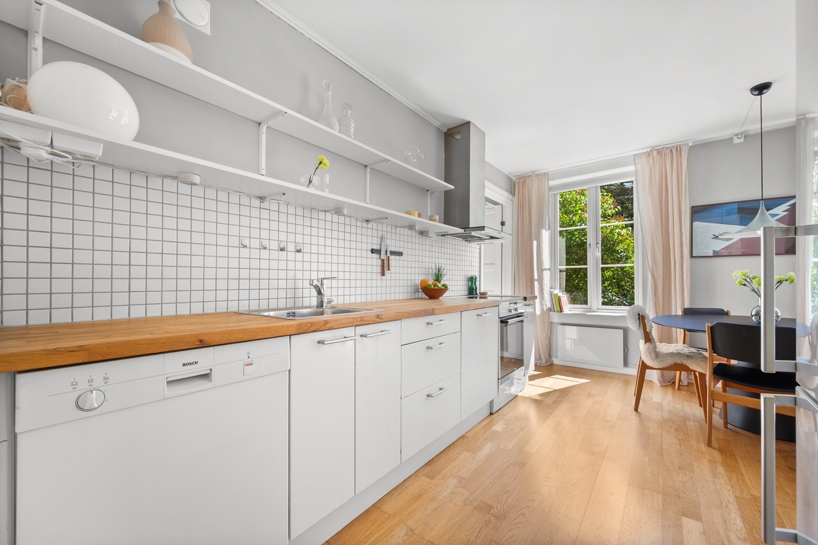 Kjøkkeninnredning med hvite, glatte fronter, fliser på vegg over kjøkkenbenk og heltre benkeplate. Galleribilde