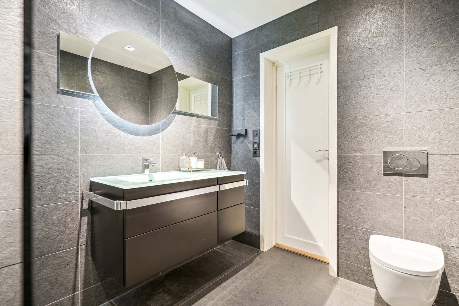 Flott baderom med mørke fliser på vegg og gulv, utstyrt med elektriske varmekabler. Høyglans baderomsinnredning og helstøpt servantplate. Galleribilde