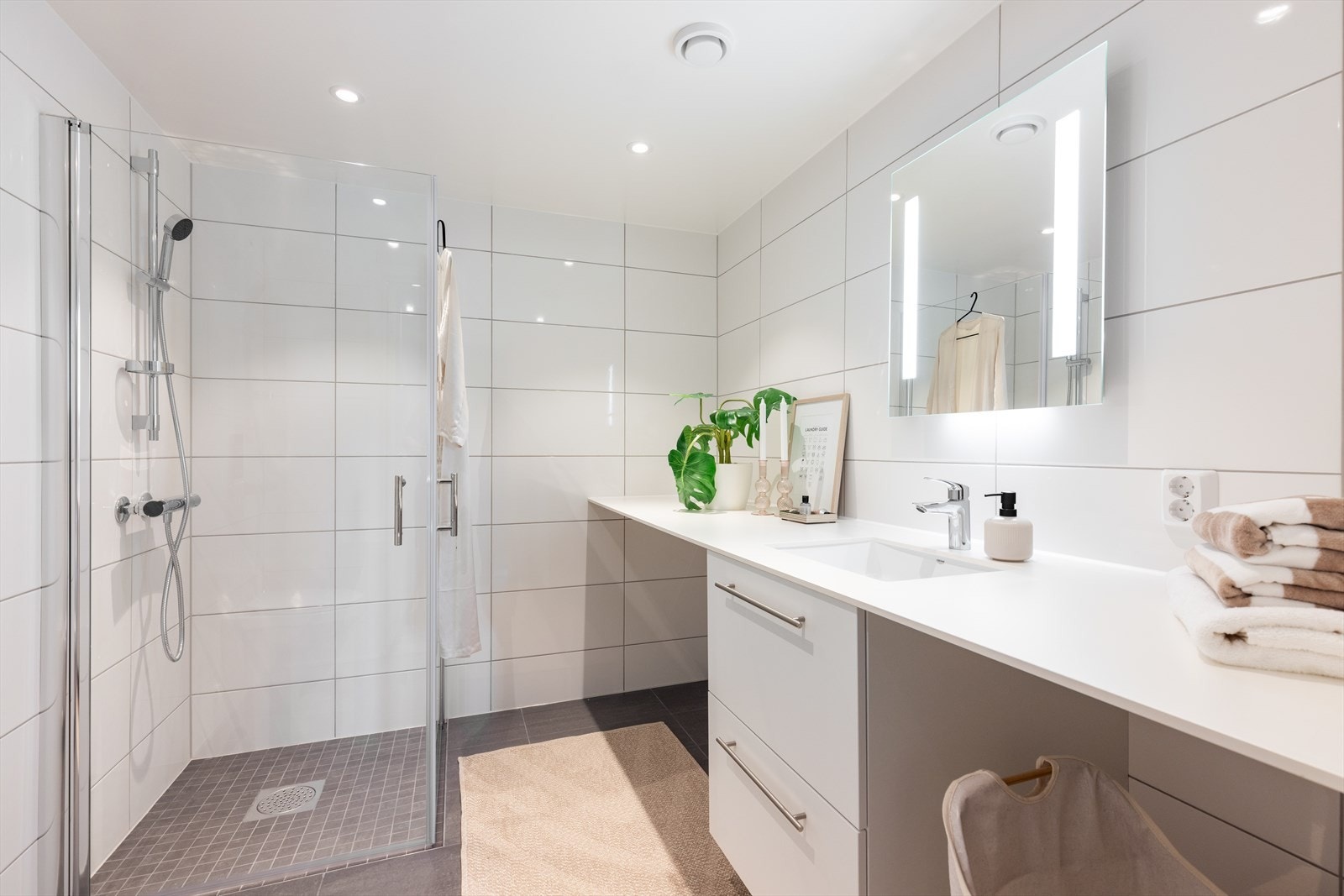 Romslig og flislagt baderom med varmekabler på gulv. Vaskemaskin og tørketrommel kan enkelt og plassbesparende plasseres under heldekkende benkeplate Galleribilde