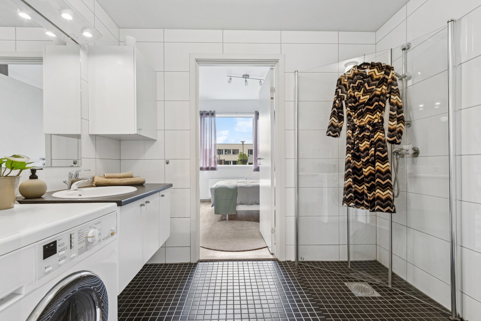 Lyst og tidløst baderom utstyrt med servantinnredning, speil med overlys, dusjhjørne, toalett og opplegg for vaskemaskin. Galleribilde
