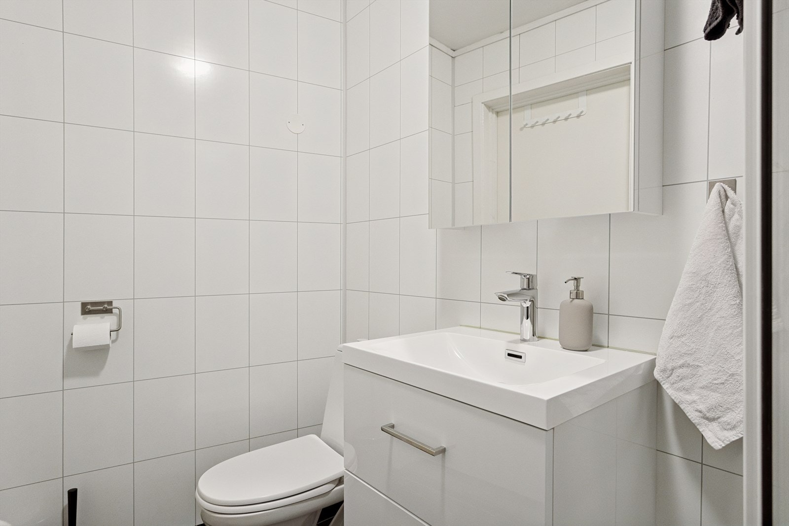 Lyst flislagt bad utstyrt med dusjkabinett, toalett, overhengt speilskap og servant. Galleribilde