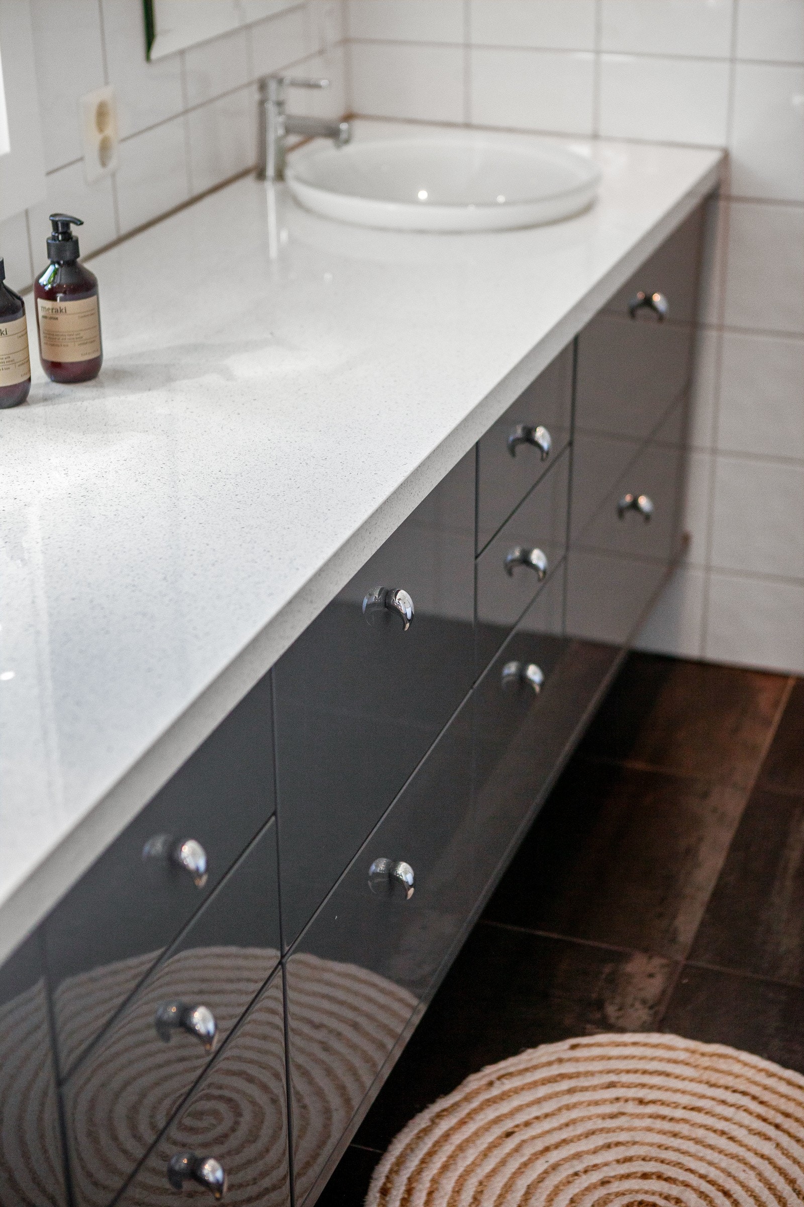 Baderomsmøblementet strekker seg langs hele ytterveggen under et vindu og har mørkegrå høyglansfronter, lys benkeplate i laminat og to nedfelte servanter. Galleribilde