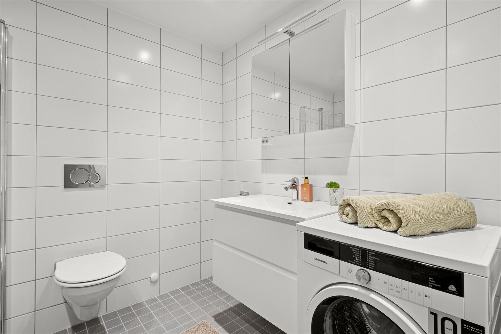 Innredningen består av overskap med speildeil og servant med hvitt underskap. Veggfestet klosett med innebygget sisterne og opplegg/plass for vaskemaskin. Galleribilde