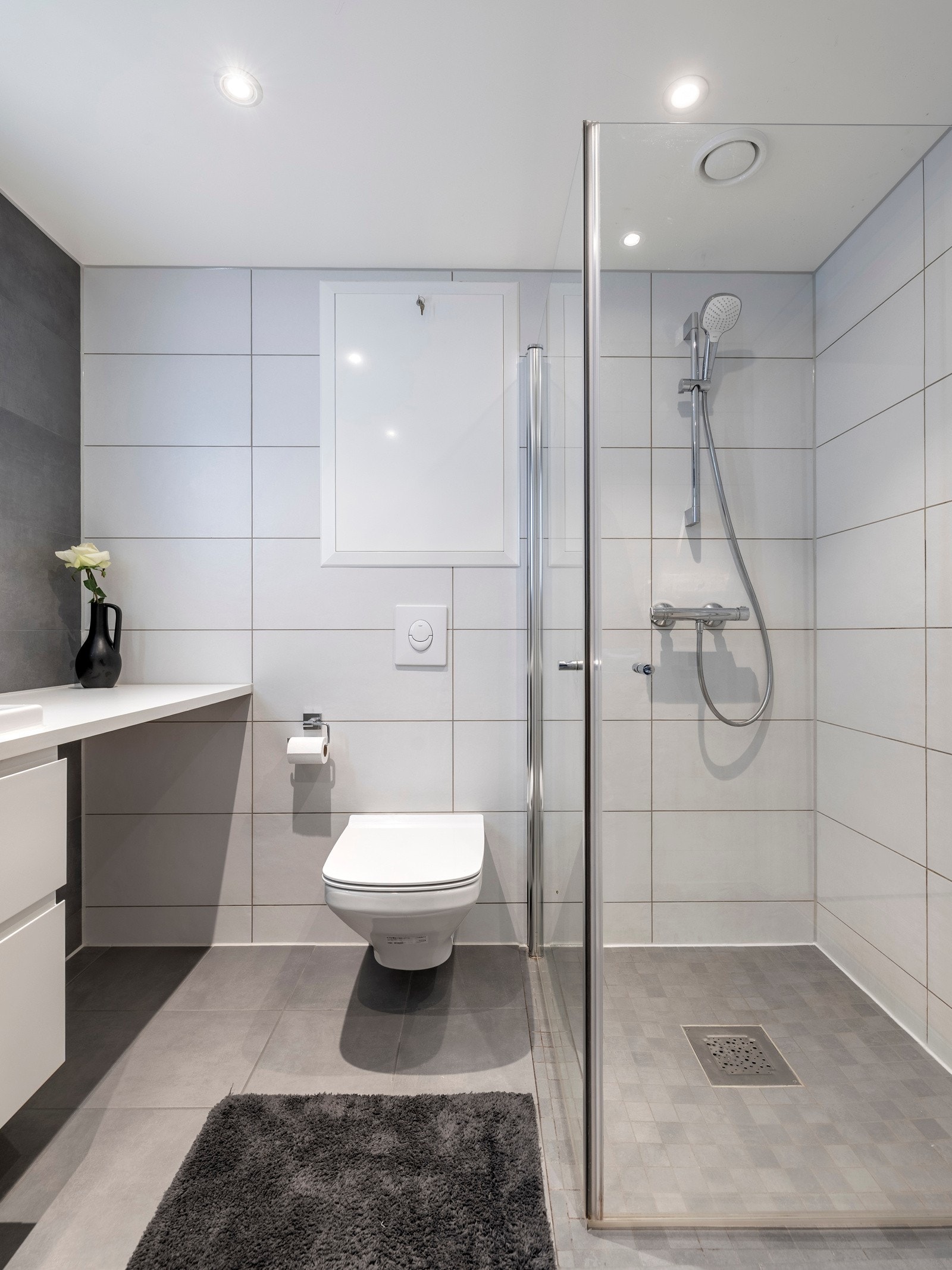 Innredningen er stilfull med en servant nedfelt i en bred plate, en hvit dobbel skuffeseksjon, et speil med integrert belysning, et veggmontert toalett og et dusjhjørne med innfellbare glassvegger Galleribilde