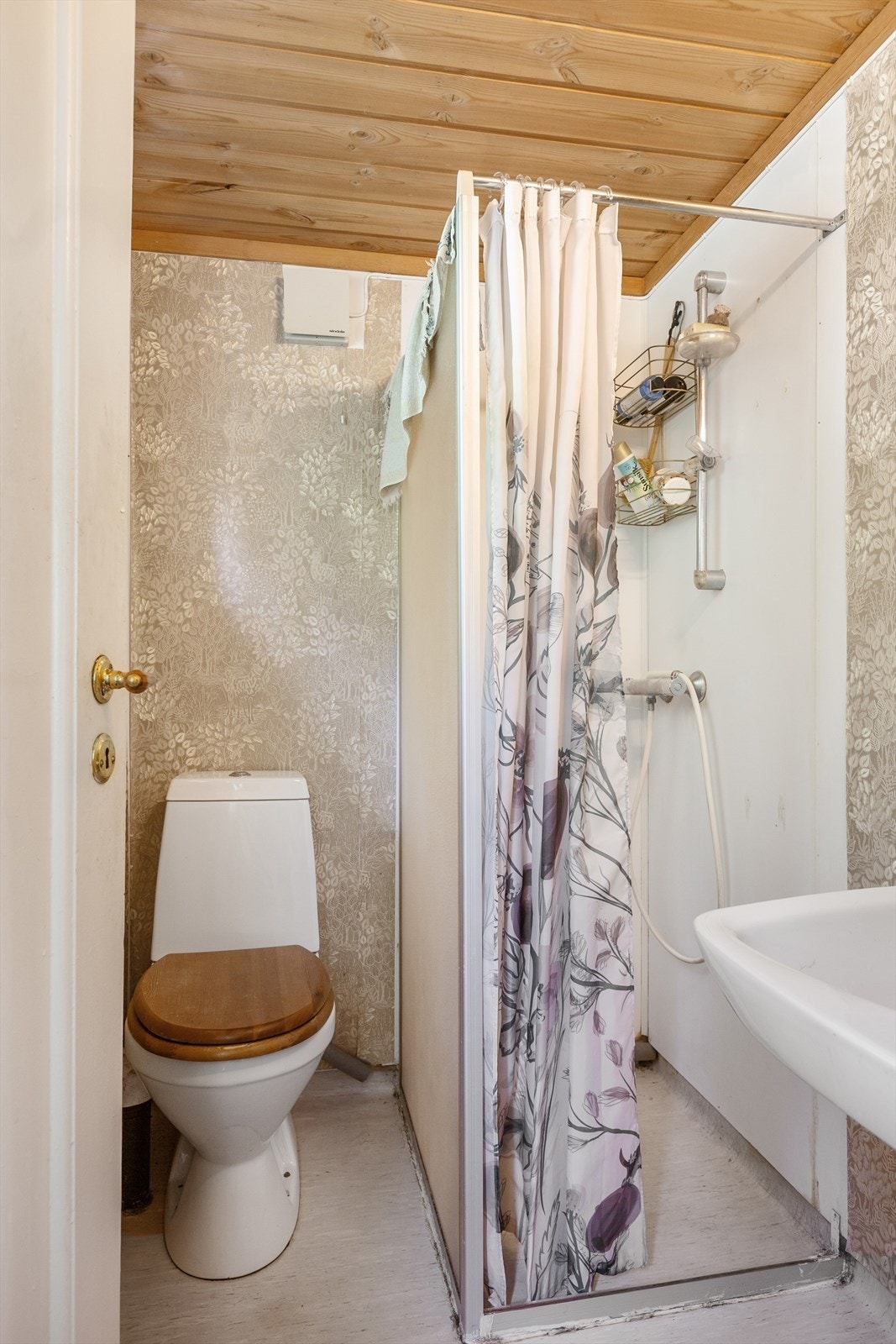 Enkelt bad med vinylbelegg, tapet og våtromsplater i dusjsone, samt servant, toalett og dusjvegg med forheng. Galleribilde