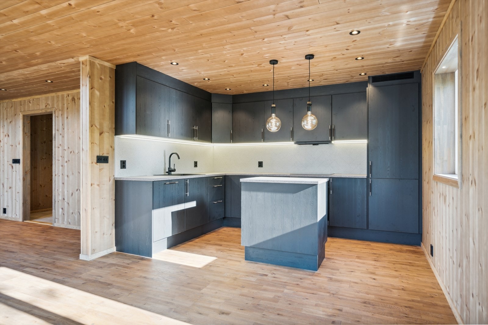 Kjøkkeninnredning med slette laminerte fronter, kjøkkenøy mot stuedel. kitchenboard over benk, nedfelt komposittvask, integrert komfyr, koketopp, oppvaskmaskn og kjøl/frys. Galleribilde