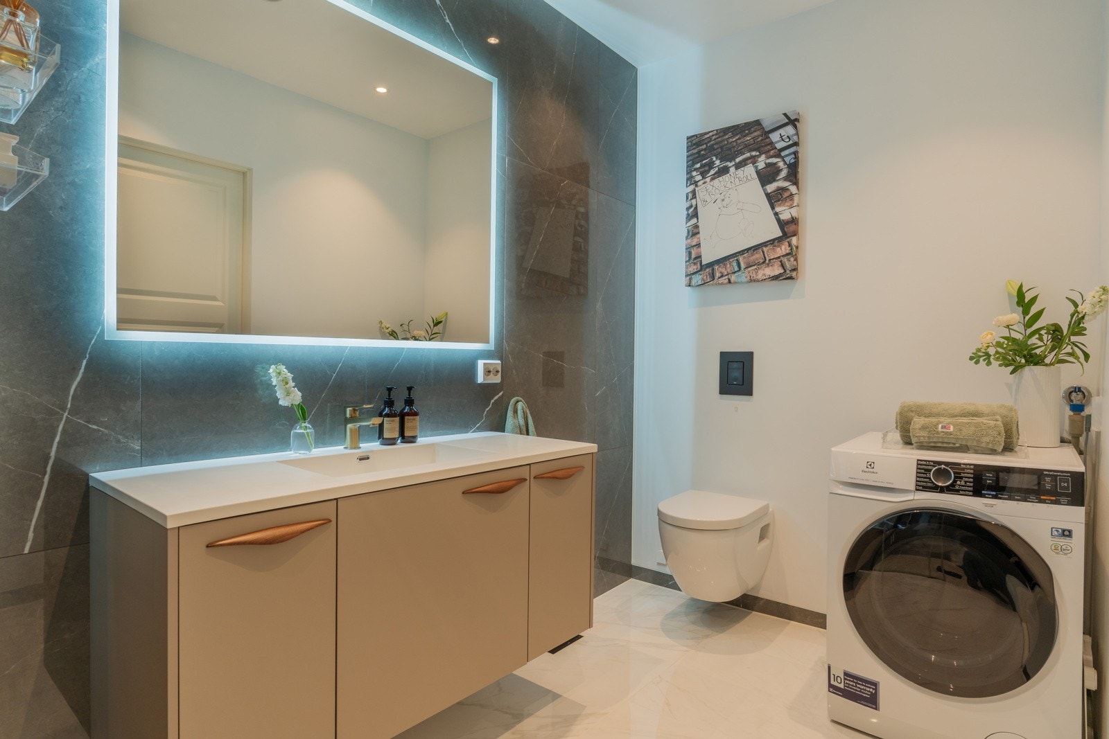 Det er gulvvarme, og innredningen består av et bredt servantskap med speil og belysning, vegghengt toalett, samt et nedsenket dusjhjørne med skyvedører og regnfallsdusj. Det er opplegg for vaskemaskin. Galleribilde