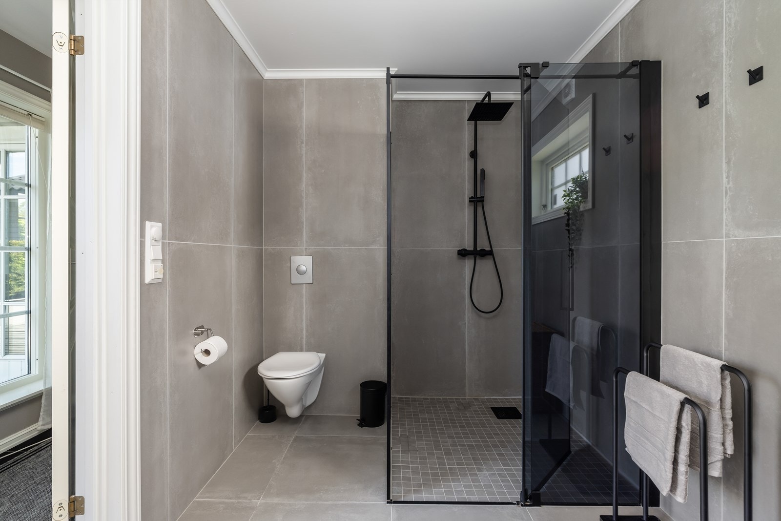 Badet inneholder veggfestet toalett, åpen dusjløsning med dusjdører og servantinnredning med underskuffer, speil og overlys. Samt samleskap rør i rør. Galleribilde