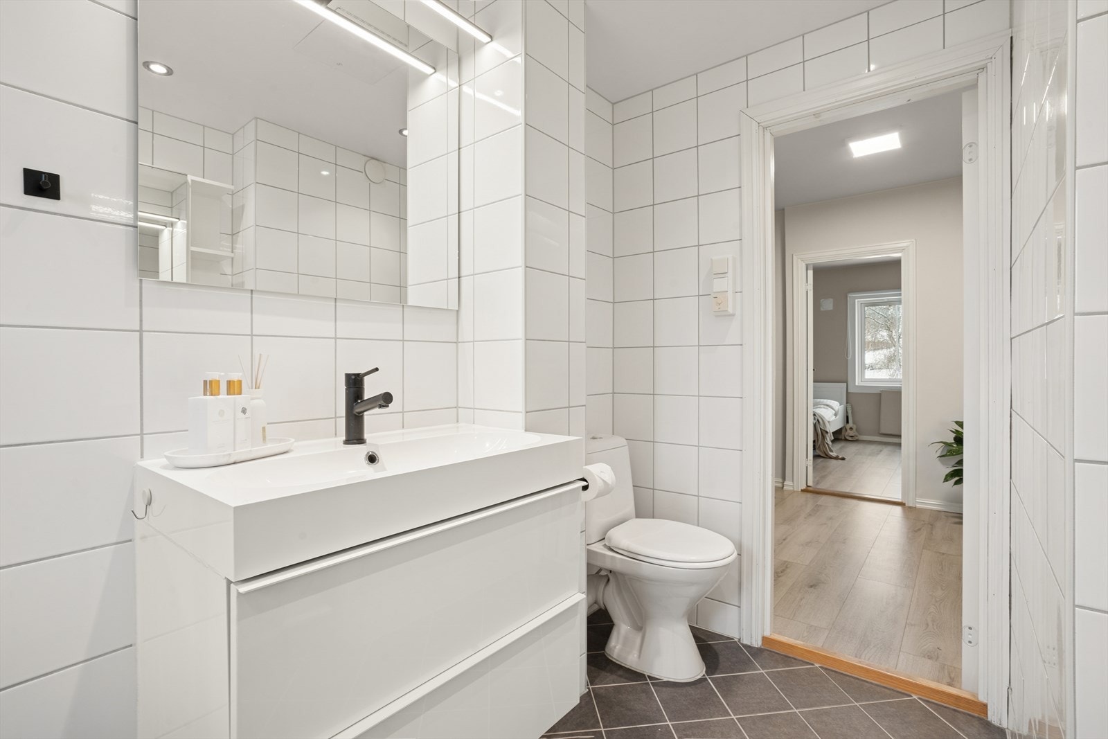 Innredning på badet består av gulvstående toalett, servant med skuffer under og speil over servant med lysarmatur. Badet har varmekabler i gulvet. Galleribilde