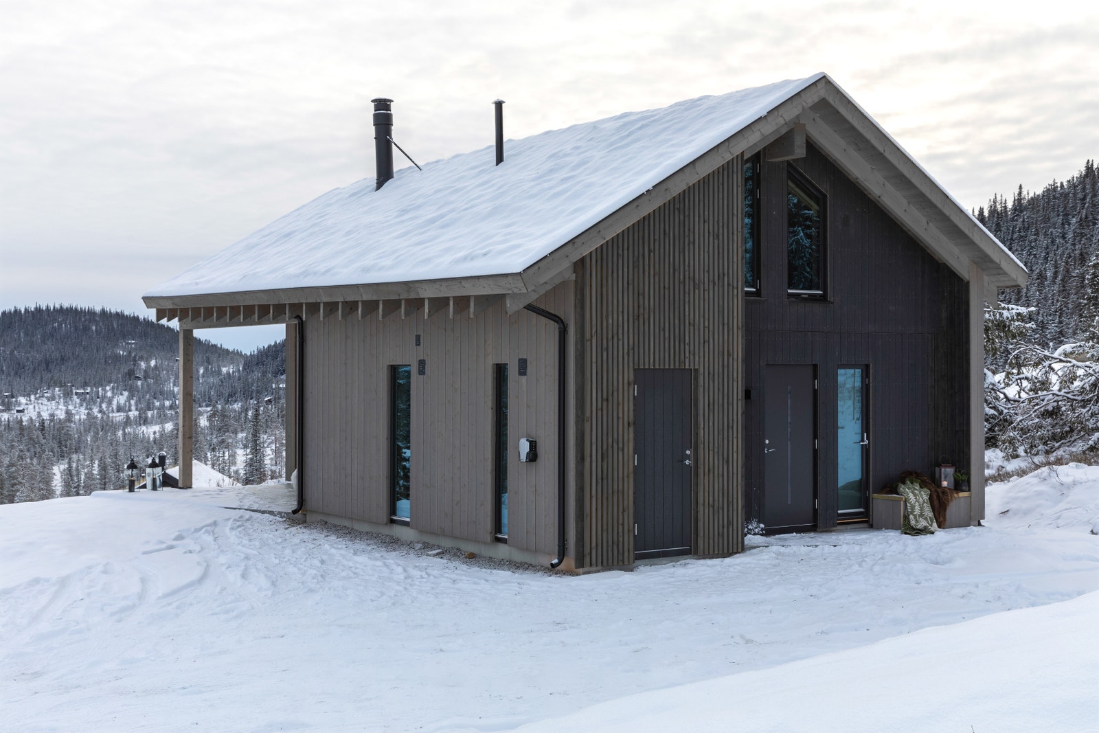Ved inngangspartiet er det også en fastmontert benk. Her kan man slappe av etter aktiviteter. Bak døren til venstre på gavlveggen er det en praktisk skibod. Til høyre er det utgang fra badet. Stålpipa på hytta har rund avslutning over tak. Galleribilde