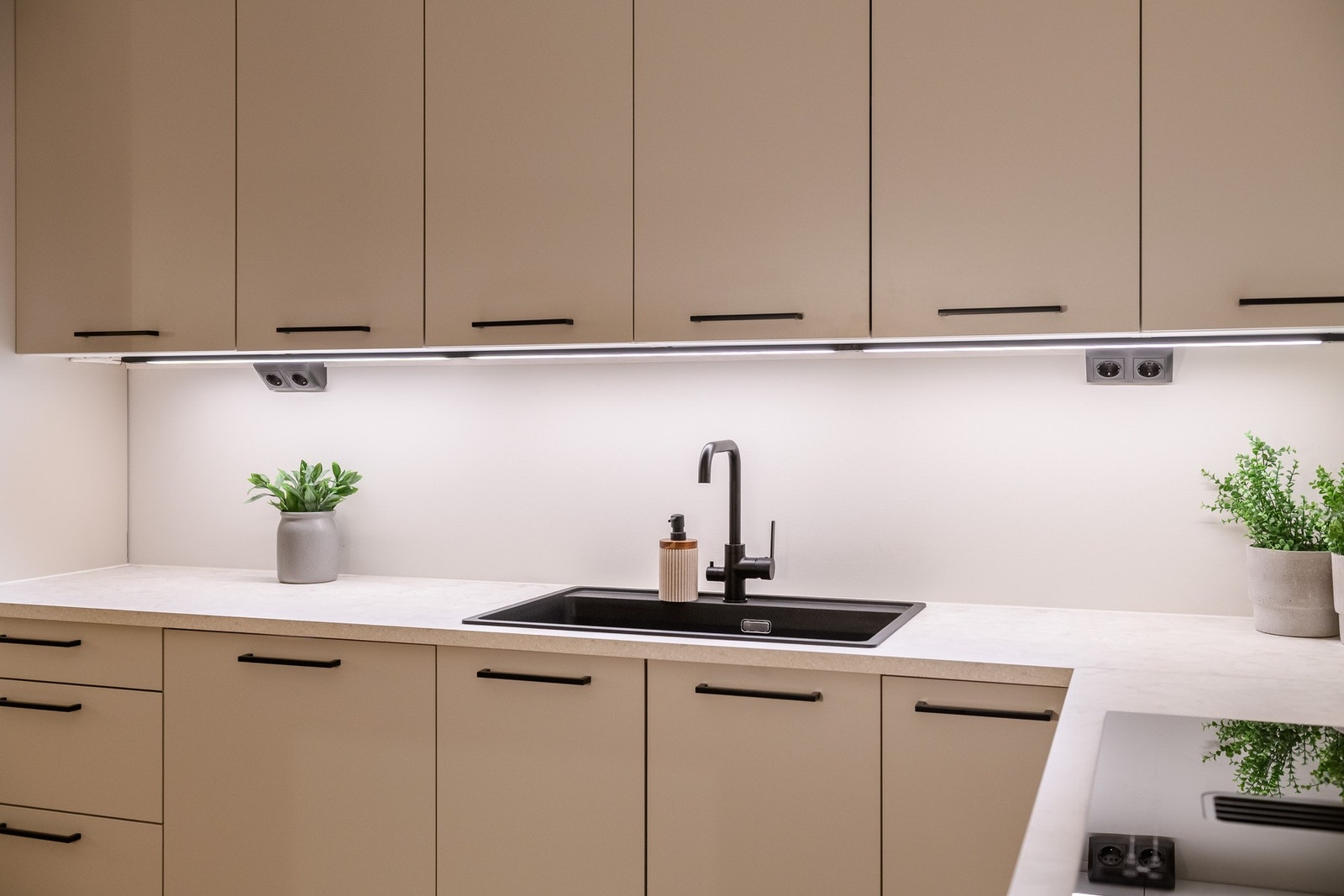 Stilig med lyse, glatte fronter og benkeplate, kombinert med håndtak, vaskekum, stikk og hvitevarer i sort design. Galleribilde