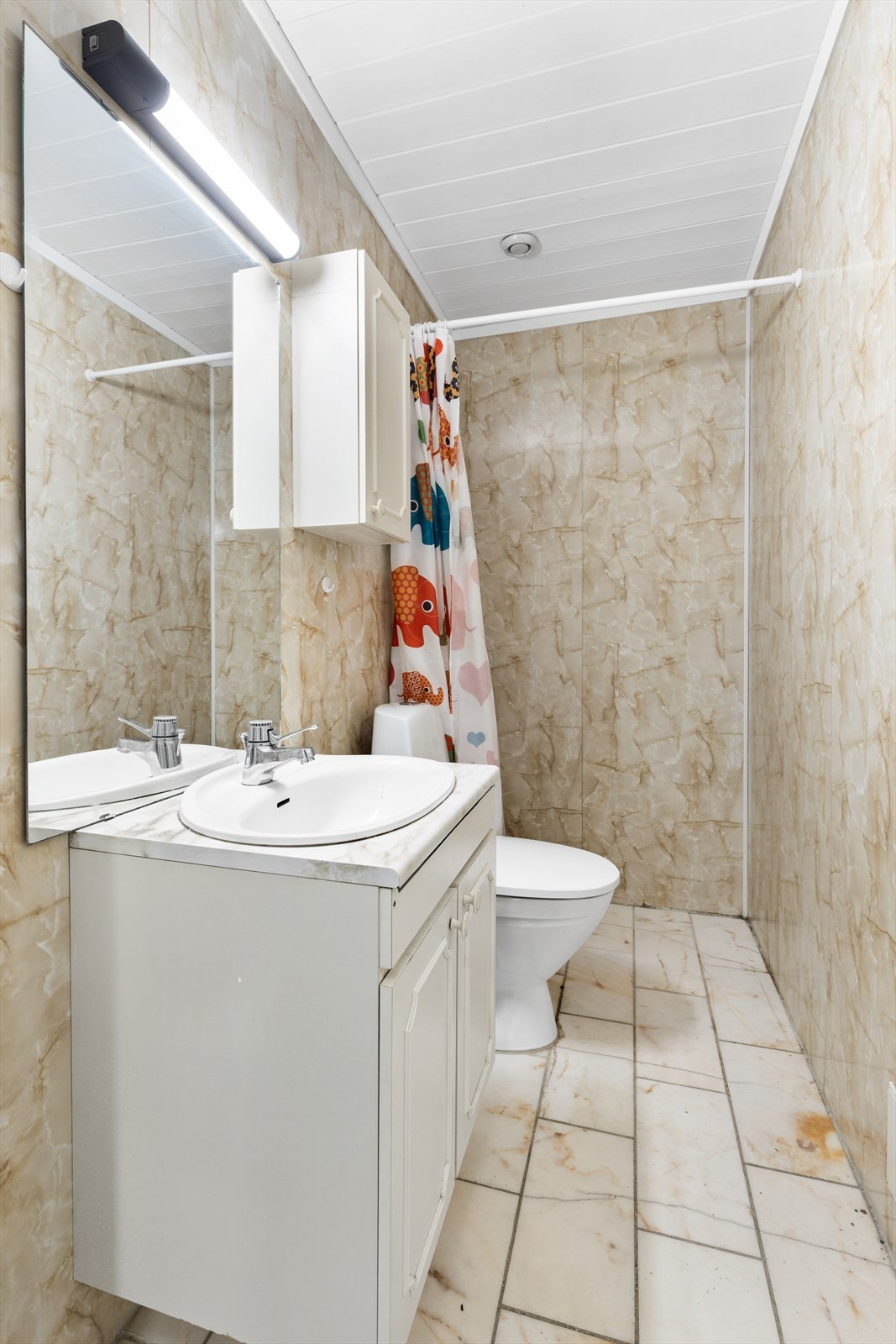 Bad fra byggeår. Flislagt gulv. Vegghengt servantskap med ovenpåliggende servant og ett-greps armatur. Speil, belysning og overskap over servant. Gulvstående toalett. Dusjsone med armatur tilkoblet hånddusj. Avtrekksventil i himling. Galleribilde