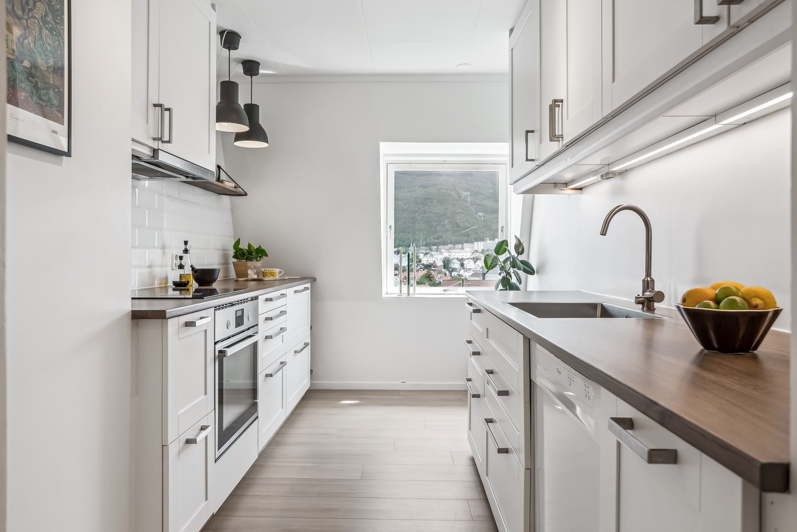 Ikea kjøkken med hvite profilerte fronter og fliser på vegg mellom over og underskap. Benkeplaten er i laminat med nedfelt stålvask. Det er integrert komfyr og induksjonstopp, samt opplegg for oppvaskmaskin og plass til kjøleskap Galleribilde