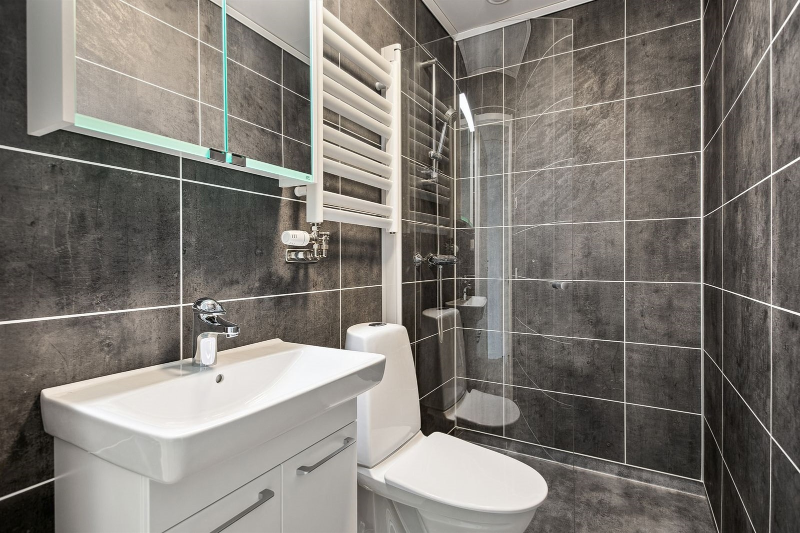 Bad nr.2 består av enkelservant, gulvstående toalett, dusjhjørne og håndkletørker over toalettet. Som på det andre badet, er det også mørke fliser på vegg og gulv. Samt komfort - varmekabler. Galleribilde