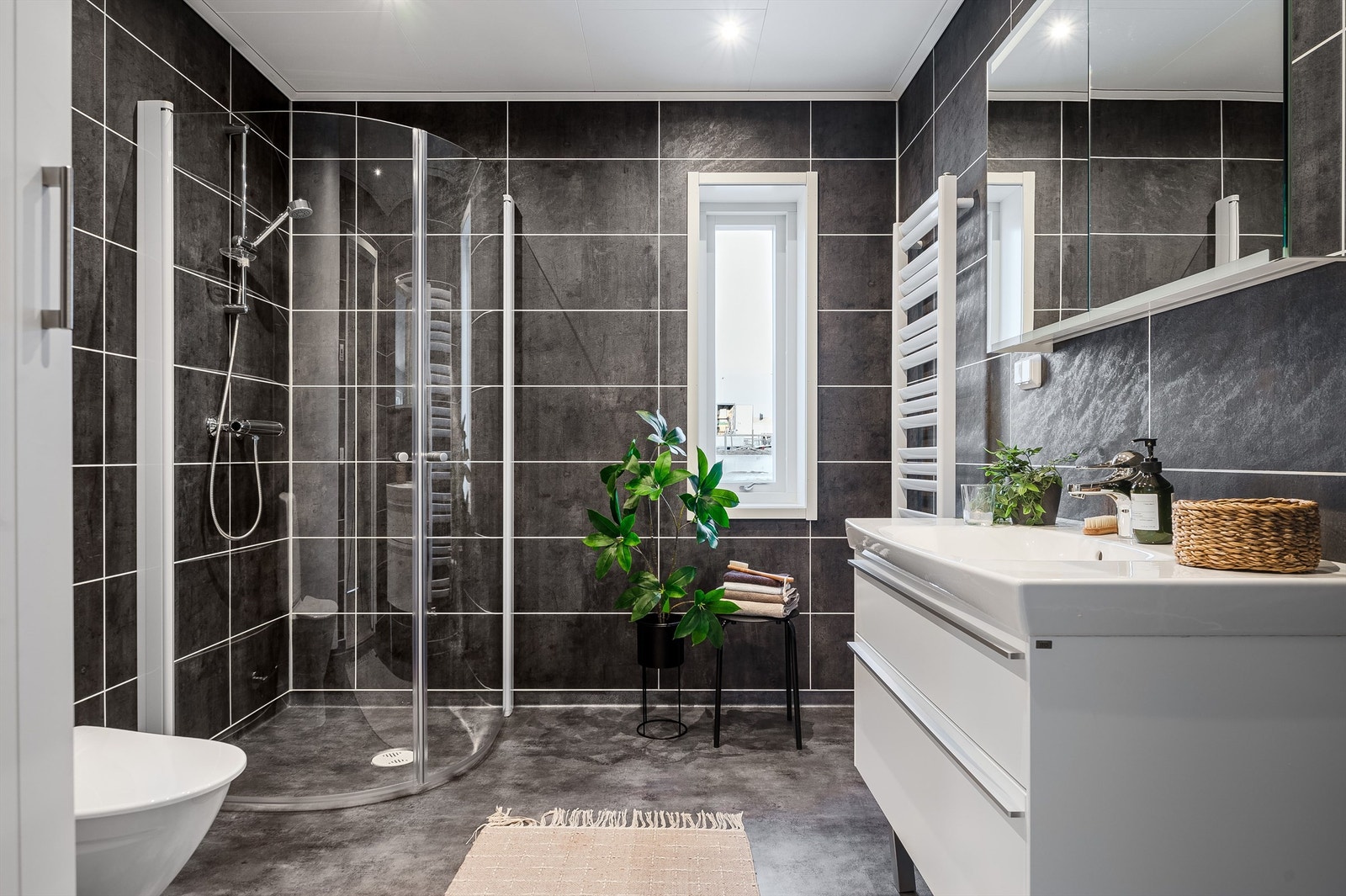 Bad nr.1 er av god størrelse. Her er det mørke detaljer på vegger og gulv som skaper god atmosfære. Badet er bestående av gulvstående toalett, enkelservant, speil med belysning, dusjhjørnet med innfellbare glassdør og håndkletørker. Galleribilde