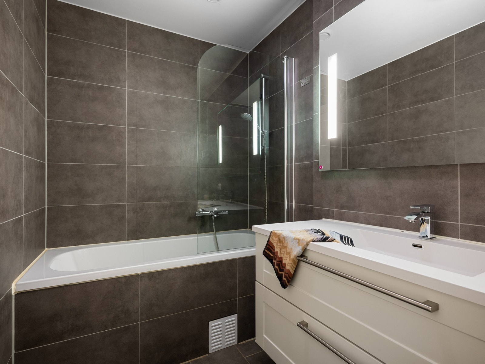 Badet er innredet med seksjon med vask og skuffer, speil med lys, vegghengt wc og badekar med dusj Galleribilde