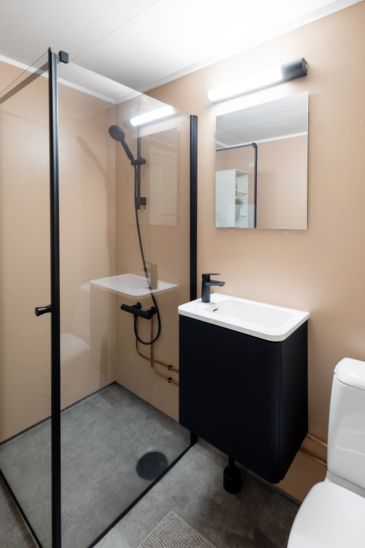 Badet er praktisk innredet med et dusjhjørne med rette, innsvingbare glassdører, et gulvmontert toalett, og en heldekkende servant med underskap og veggmontert speil. Toalett, servant og dusj er nyinstallert. Galleribilde