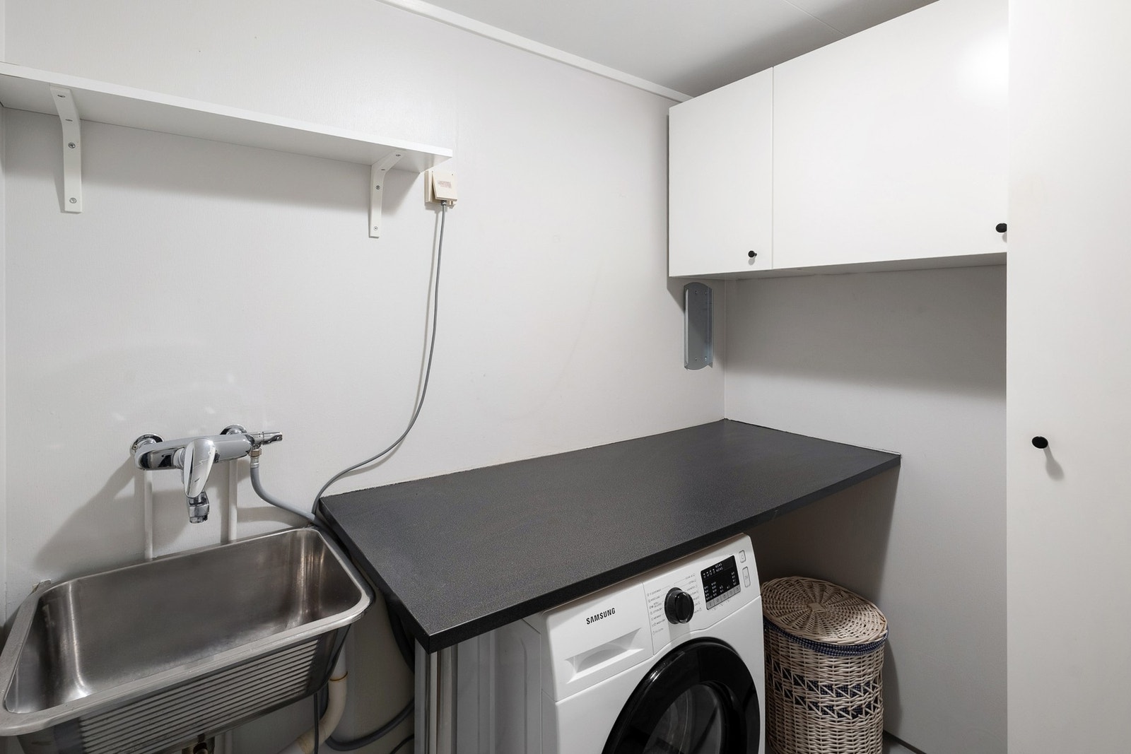 Rommet er utstyrt med stål utslagsvask og opplegg for vaskemaskin, samt praktiske skapreoler på vegg for oppbevaring. Galleribilde