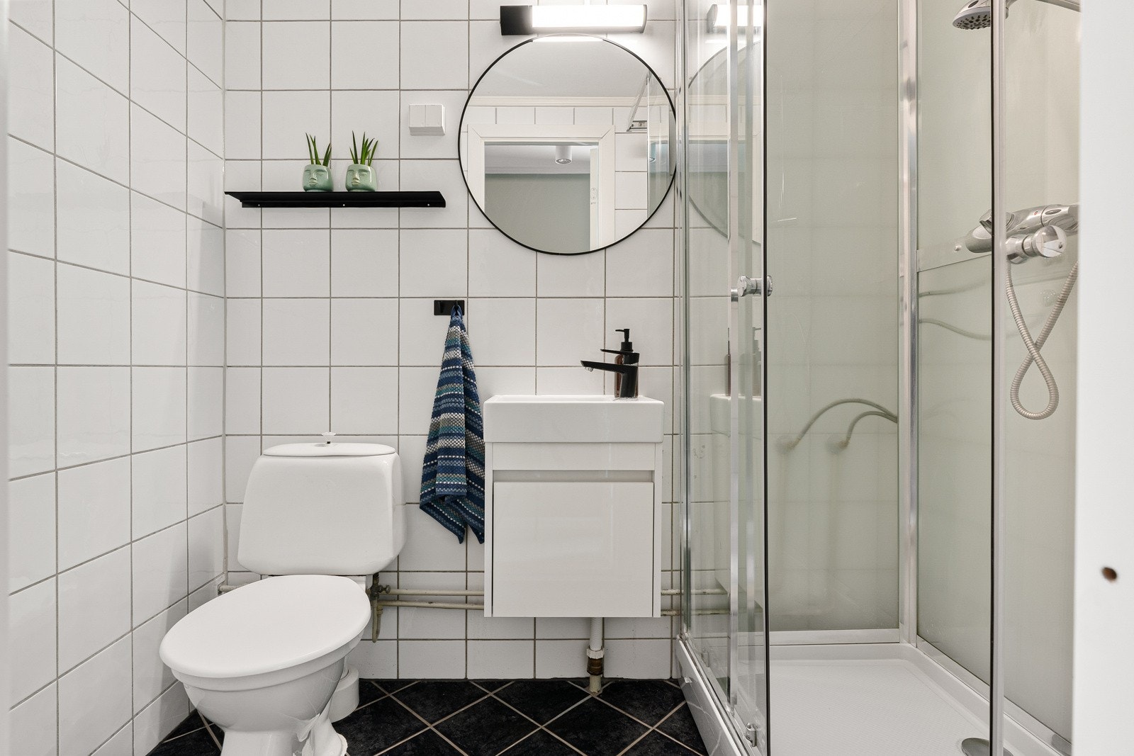 Herfra er det adkomst til leilighetens bad. Badet har flislagte overflater, gulvvarme, toalett, dusjkabinett og servant med tilhørende servantskap. Galleribilde